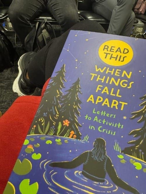 Copy of ‘Read This’ on a Atena’s lap above a dark, carpeted airport floor. The book's cover features art by Ollie Costello, a night scene with a person in a lily pond.