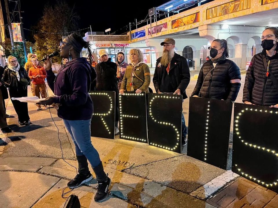 Photo: Kelly Hayes - Atena addresses a crowd in Rogers Park, sharing a poem; people hold boards that spell RESIST ICE in lights behind her.
