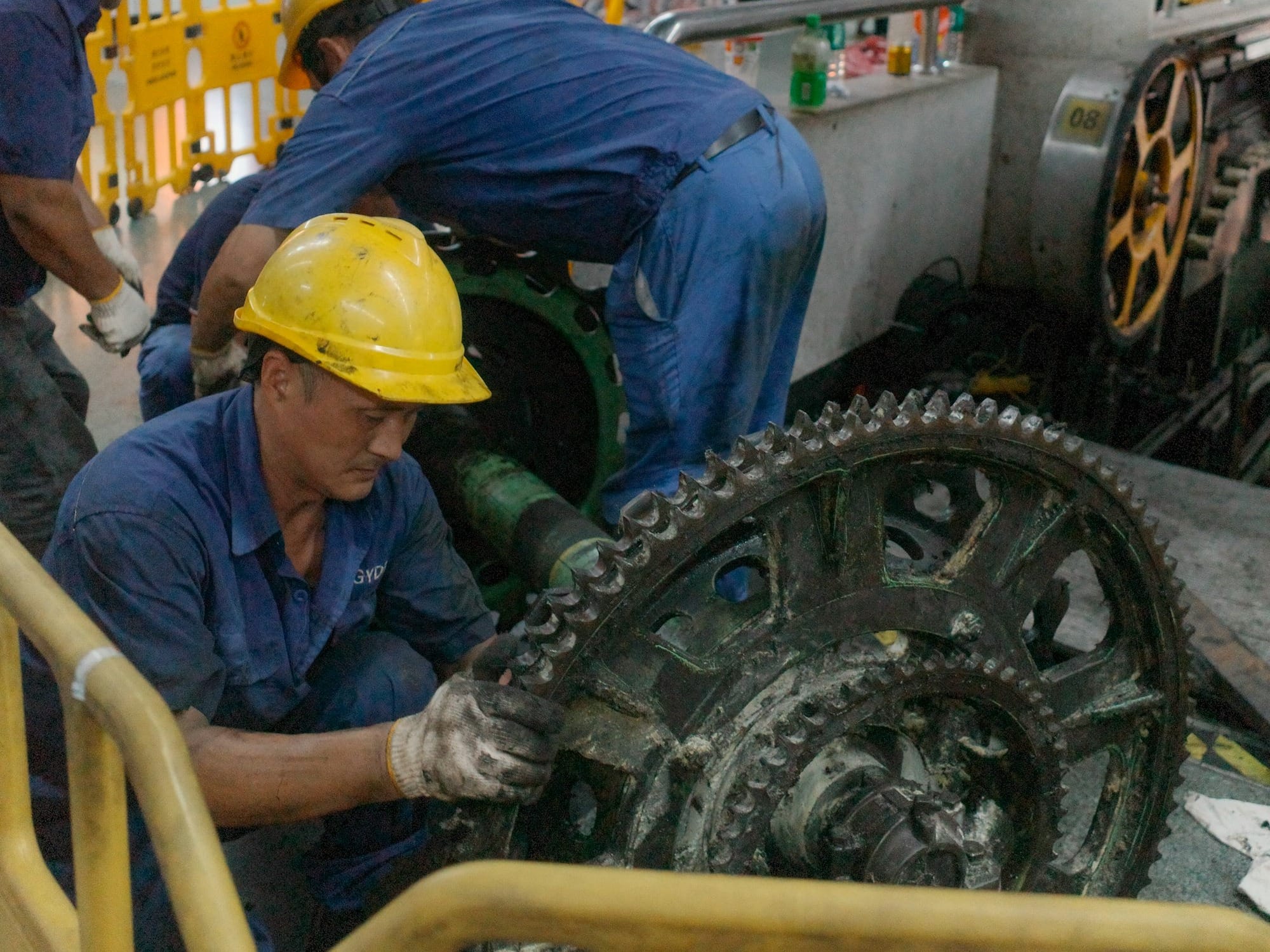 Workers in hard hats repair large industrial machinery gears.
