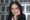 Arundhati Roy rests her chin on her hand and looks directly at the camera. She is seated indoors in front of shelves filled w