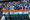 A crowd of Indian cricket fans, many in blue jerseys, behind a row of three Indian flags in the stands during the first Test 
