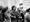 A black and white image of a group of protesters walking side by side with arms interlinked. One protester is carrying a post