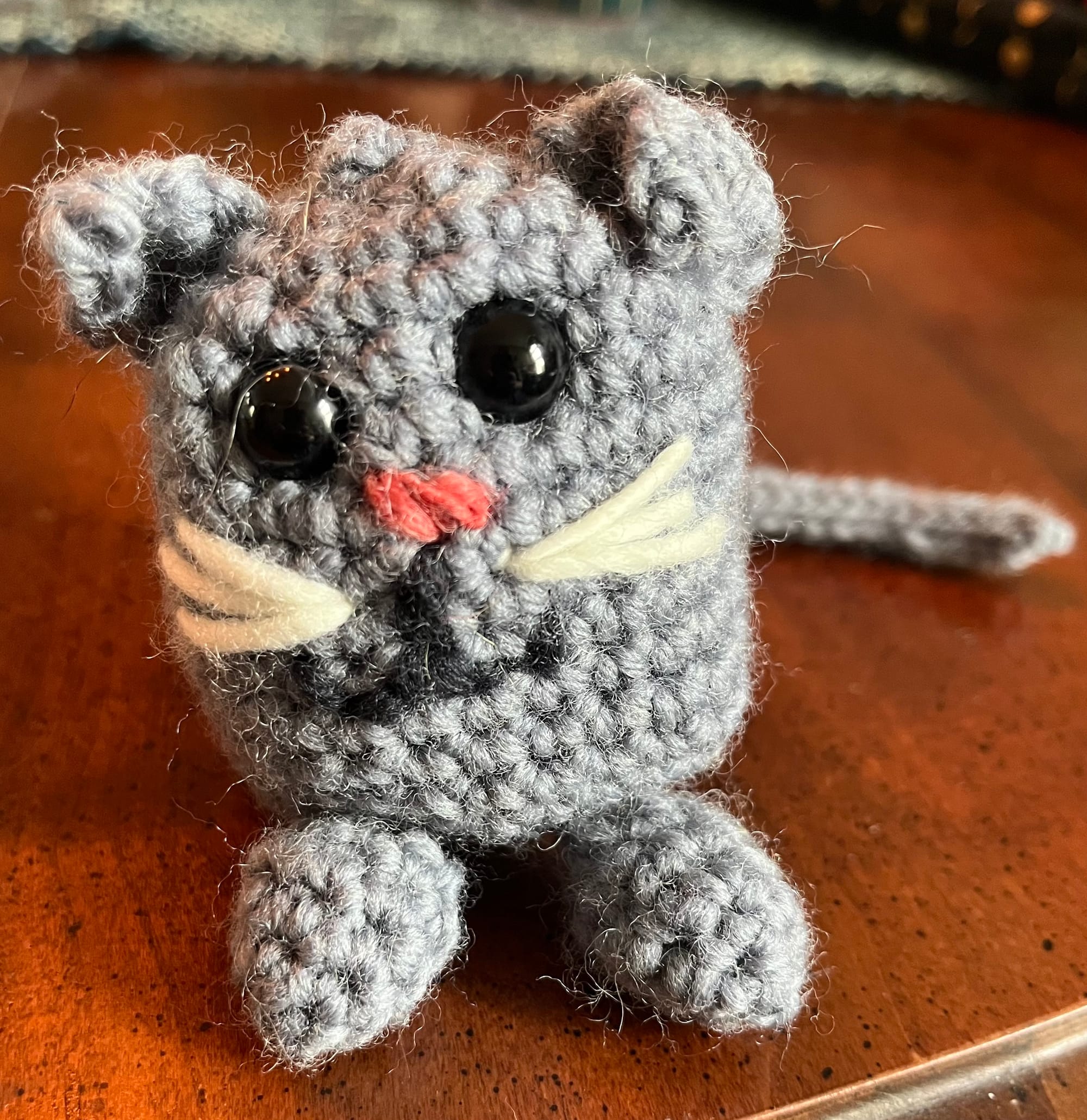 A spherical work of crochet representing a small cat. Her face is embroidered on with black and white yarn, and she has shiny plastic safety eyes. Her ears are slightly folded, representing the Palestinian Fold's distinctive look. A cute lil tail is poking from around her left side, and she rests on a cherry wood colored table.