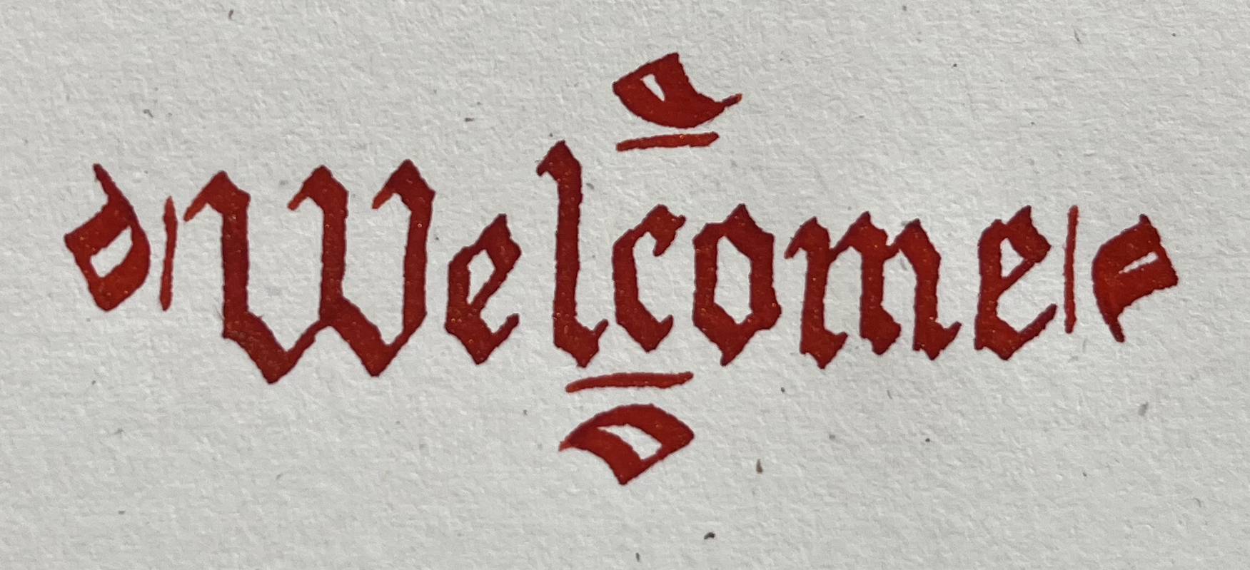 The English word "welcome" in red gothic letters, hand inked on a textured, cream colored paper.