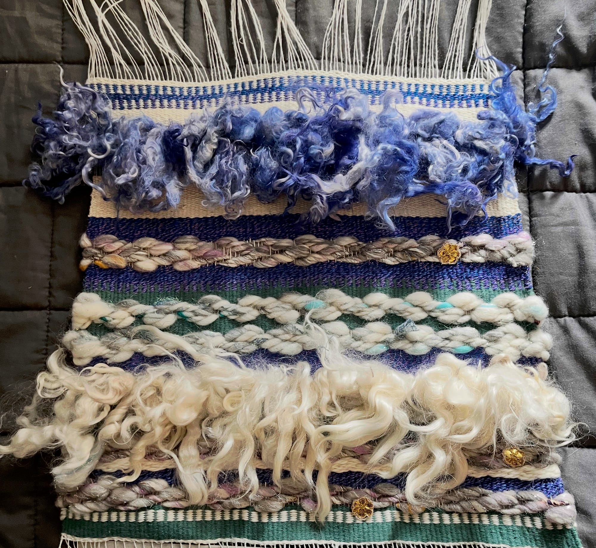 A medium sized tapestry laid flat on a grey blanket. It is woven with contrasting textures and colors in a purple, white, and green color palette.