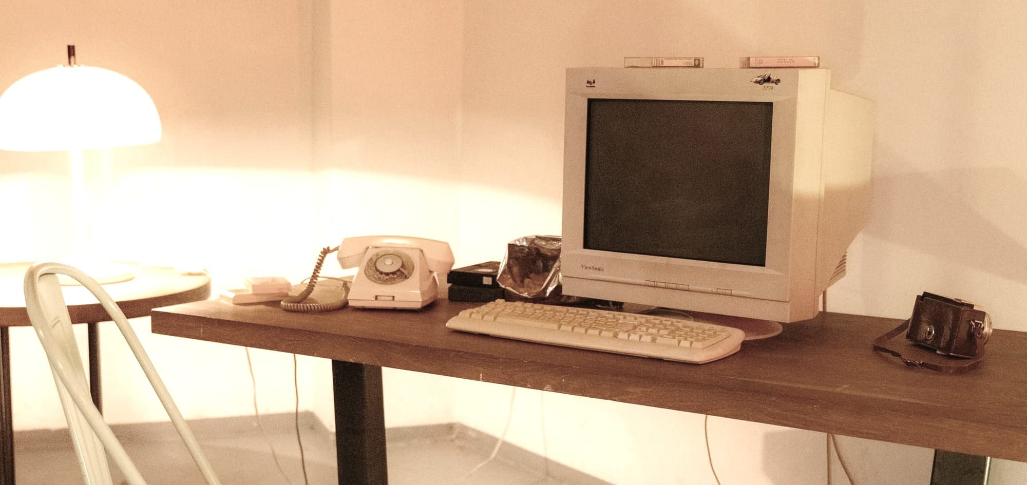 A vintage desk setup featuring a bulky CRT monitor, a rotary dial telephone, and a metal chair, all bathed in warm lighting. The scene evokes nostalgia for a pre-digital era, representing a simpler, less connected time.