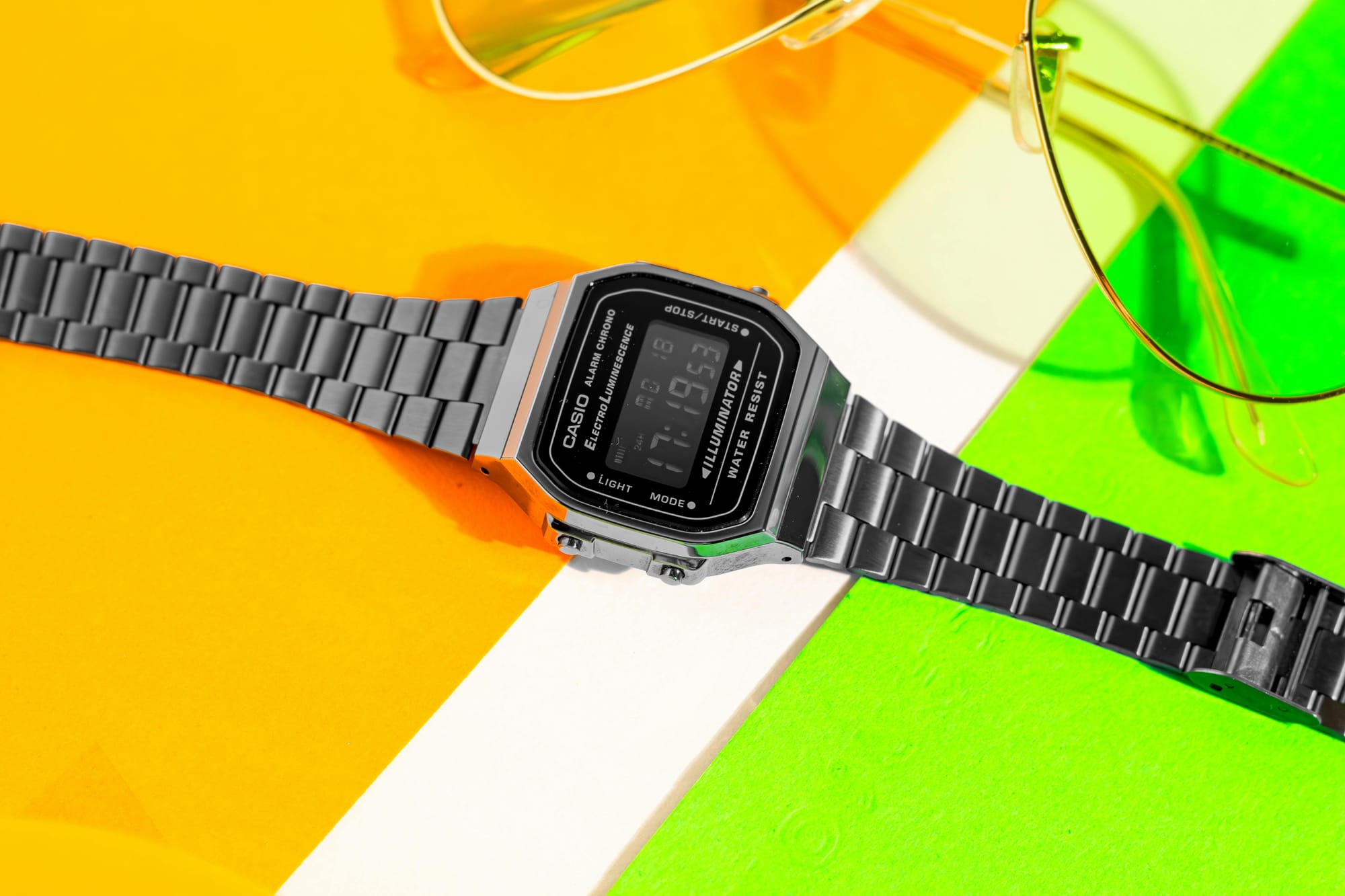 A classic Casio digital watch with a sleek metal strap, displayed on a colourful background of orange, yellow, and green, accompanied by a pair of aviator-style sunglasses. The watch symbolises a minimalist approach to technology, offering simplicity in a world of smart devices.