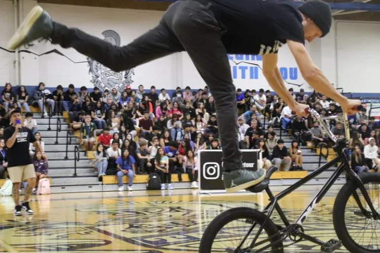Photo of a young, white man with 1 foot on his bicycle seat & the other extended as he does a trick