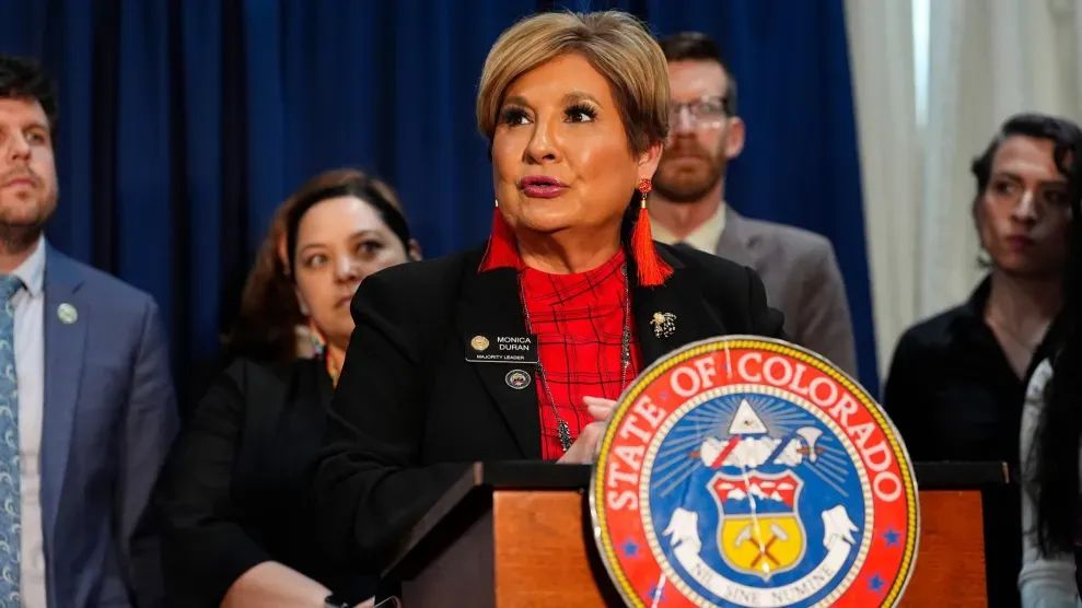 Monica Duran stand behind a lectern with the State of Colorado seal and surrounded by other people.