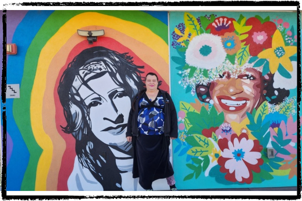 A woman w/short, black hair wearing a colorful blouse & black skirt stands in front of 2 side-by-side colorful murals of wome