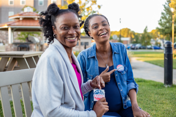 Two young, Black women sit on a park bench holding "I Voted" stickers