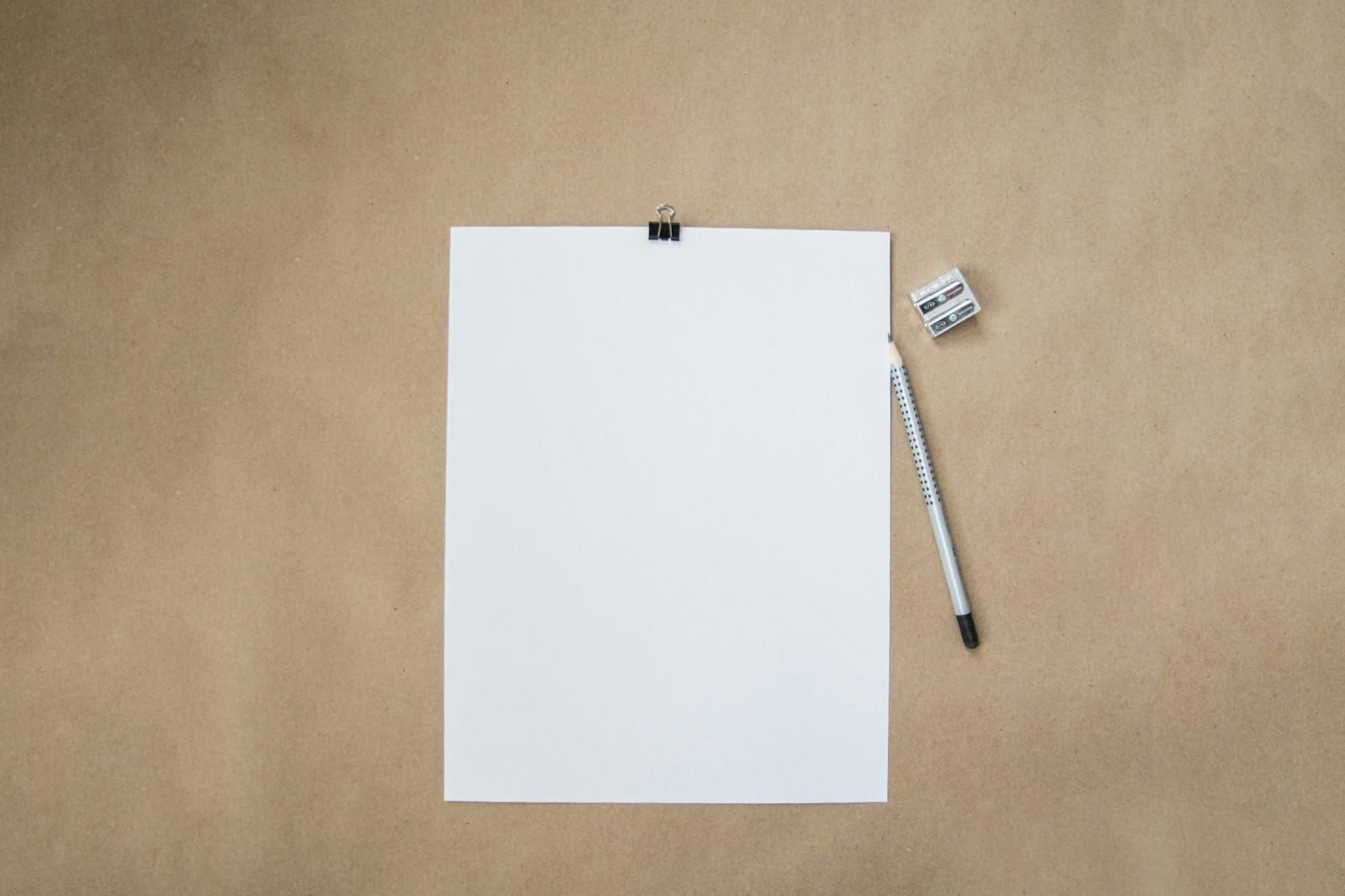 An empty sheet of white paper on a plain brown background, next to a a pencil and sharpener