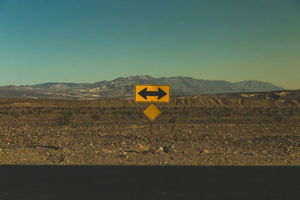 A yellow road sign with arrows pointing both left and right, against a vast, arid landscape.