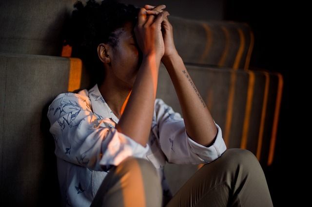 Woman leaning against wall, with hands clasped in front of her face and elbows rested on knees