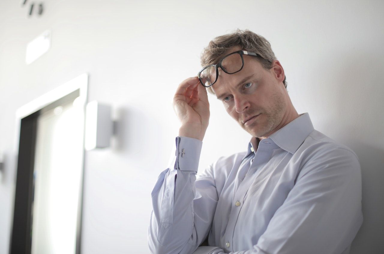 Annoyed-looking man in a shirt leaning against a wall, lifting his glasses off his face.