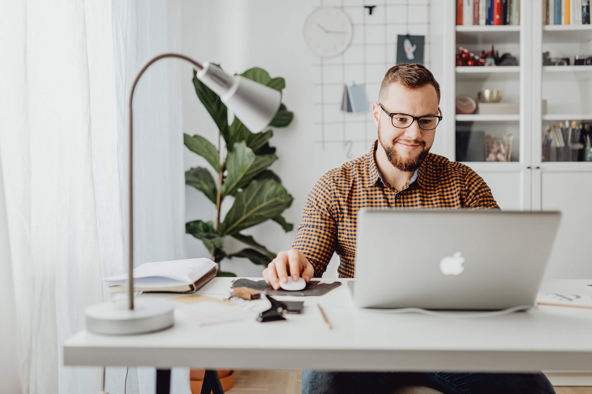 Photo of a man in checkered shirt smiling while using an Apple laptop at a desk