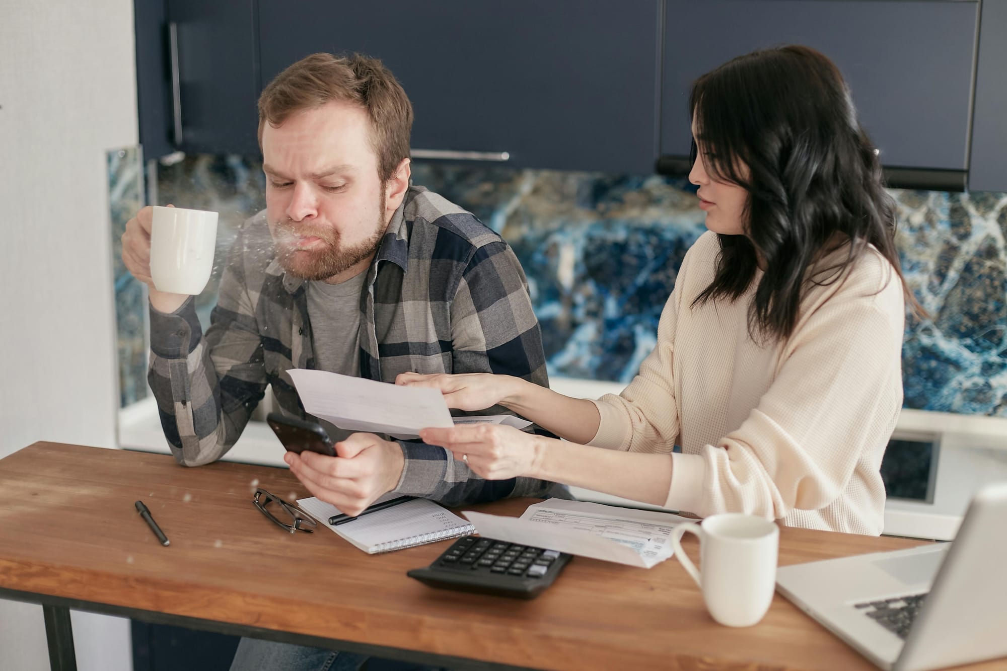 A man spitting out tea in shock as he looks upon a bill a woman shows him