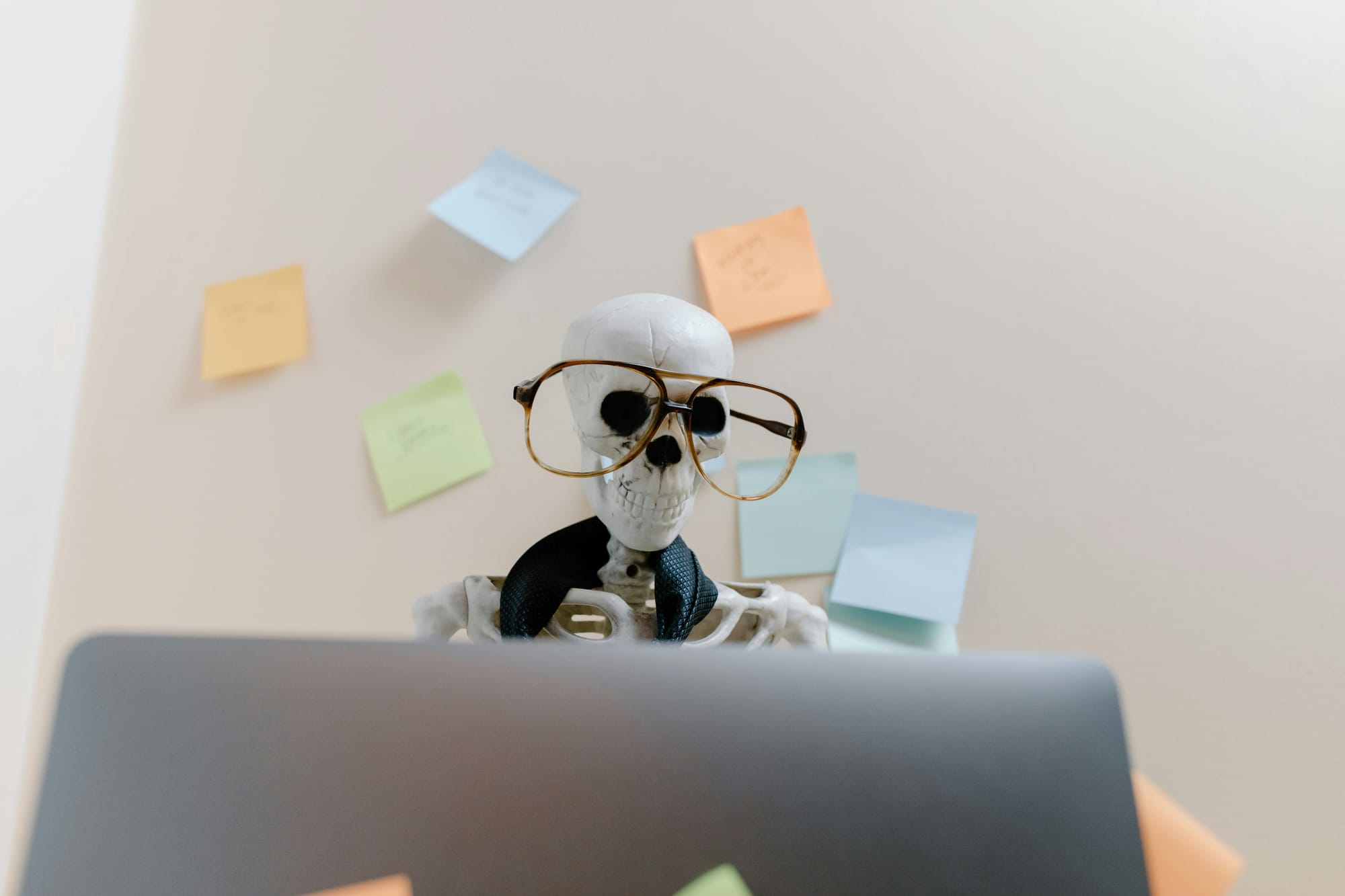 A skeleton wearing glasses and a loose tie sits in front of an open laptop with many Post-it notes plastering the wall behind it.