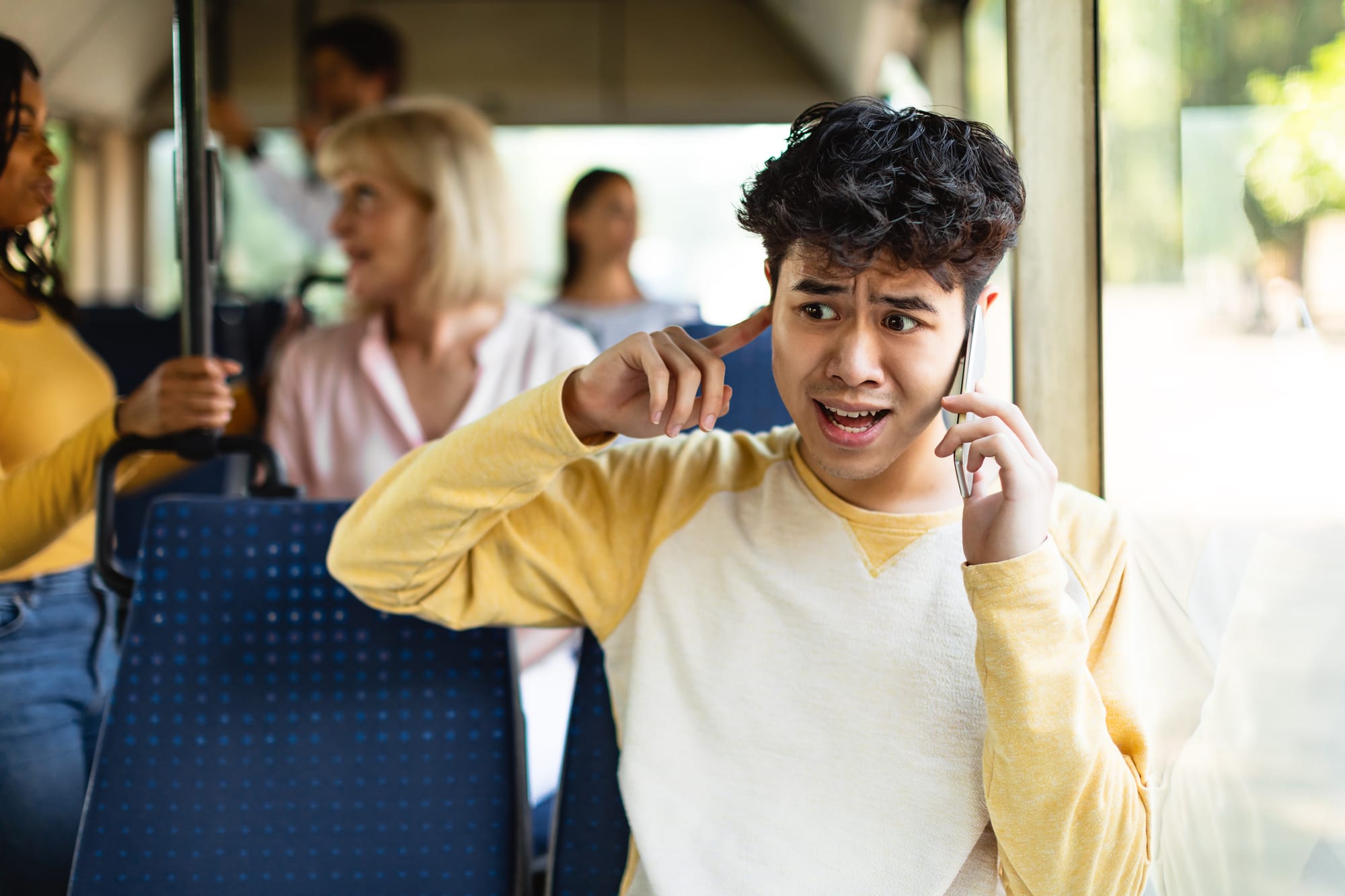A young man sits on a bus, mobile phone to one ear and a finger in the other, looking frantic.