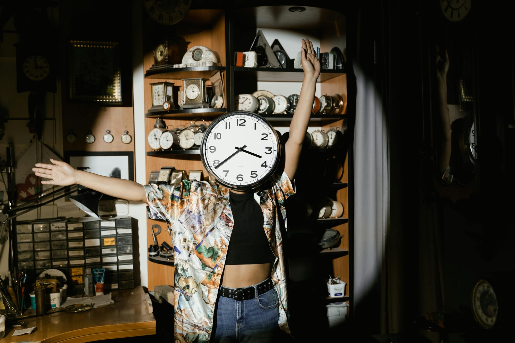 Someone with a clock for a face poses dramatically in a spotlight, shelves of clocks behind them.
