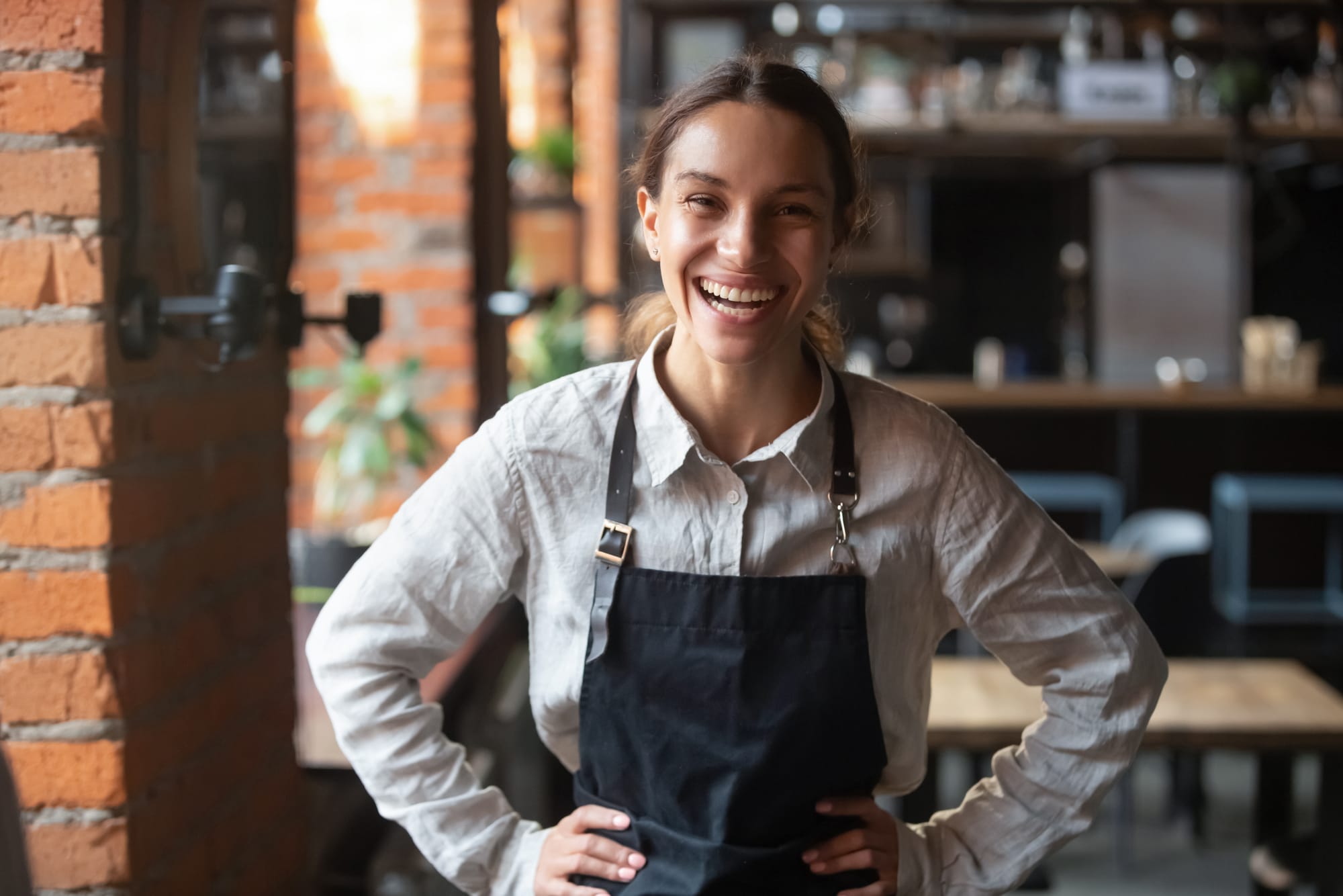 A hospitality worker in an apron, hands on hips and smiling at the camera, as if mid-laugh.