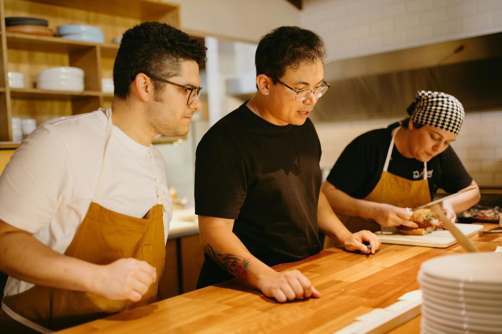Three hospitality workers. Two wear aprons and the other is dressed in all-black, the manager. The right worker preps a sandwich. The left worker listens to the manager's instructions.
