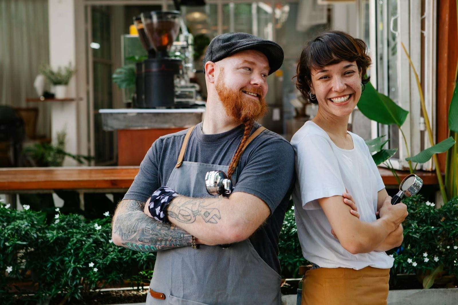 Two baristas stand back-to-back outside the cafe, one looking at the other while they smile directly at the camera.