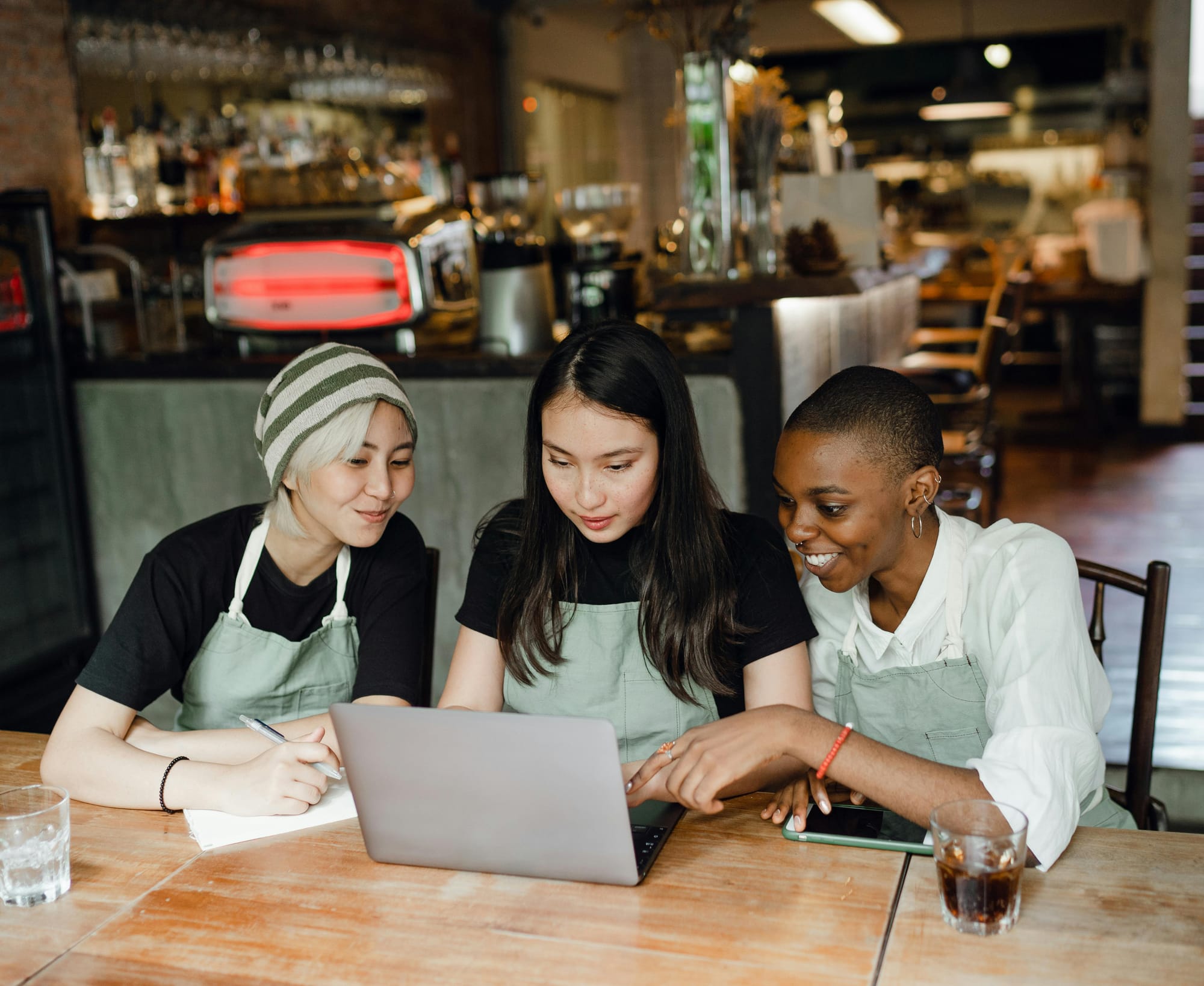 Three women in hospitality, sat around a laptop in their cafe/bar, smiling, pointing, and taking notes.
