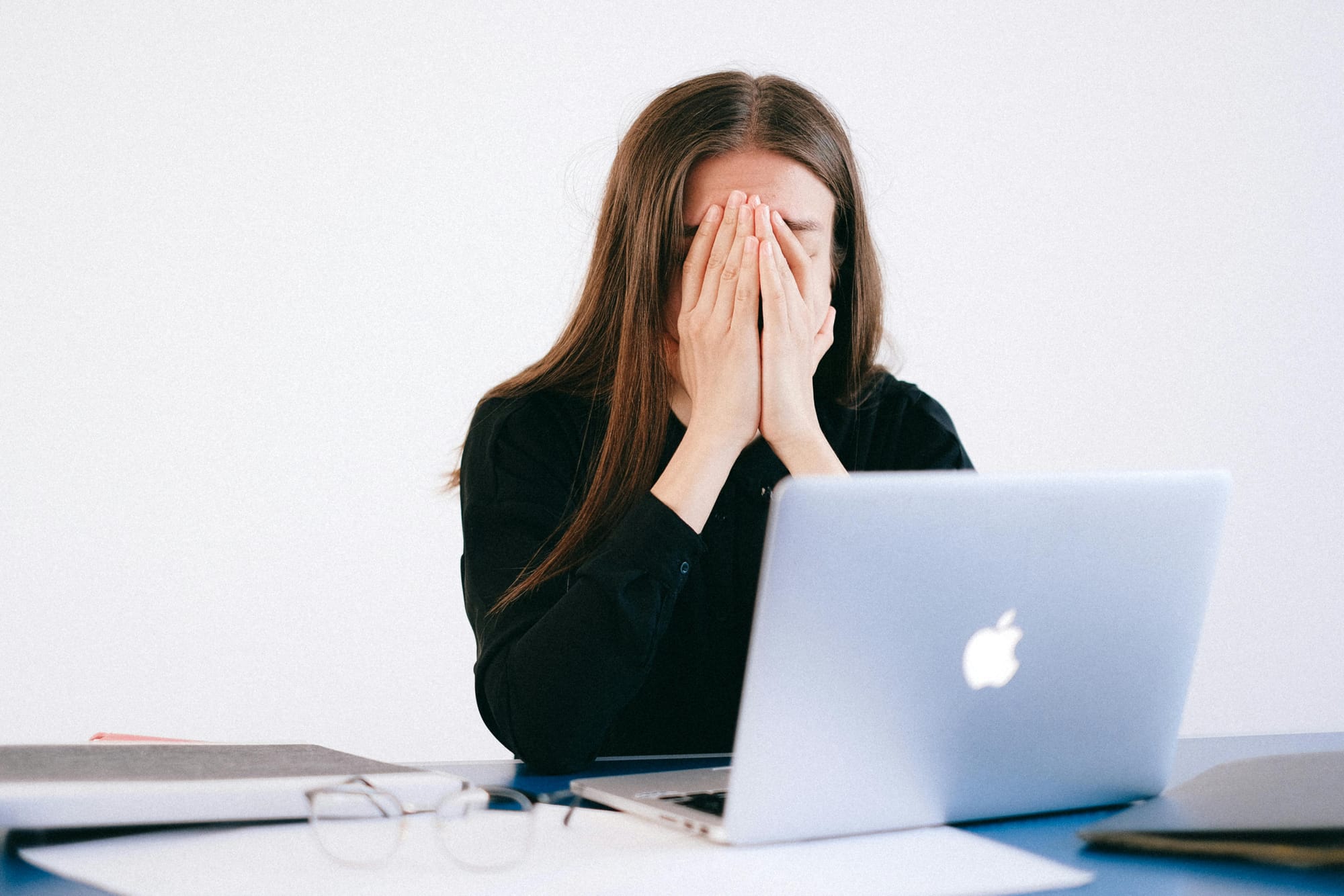 A woman at her laptop with her glasses on the table and her hands covering her face.