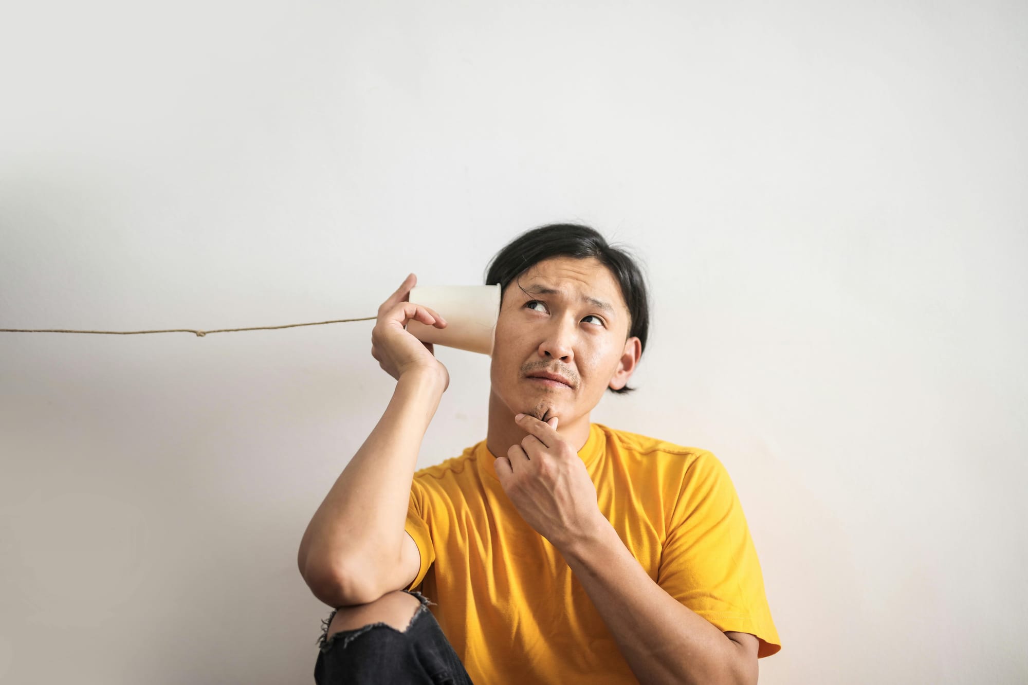 A man in a yellow t-shirt, sat against a white wall with a paper cup to his ear. A string from the cup leads away while he holds his chin, seemingly listening.