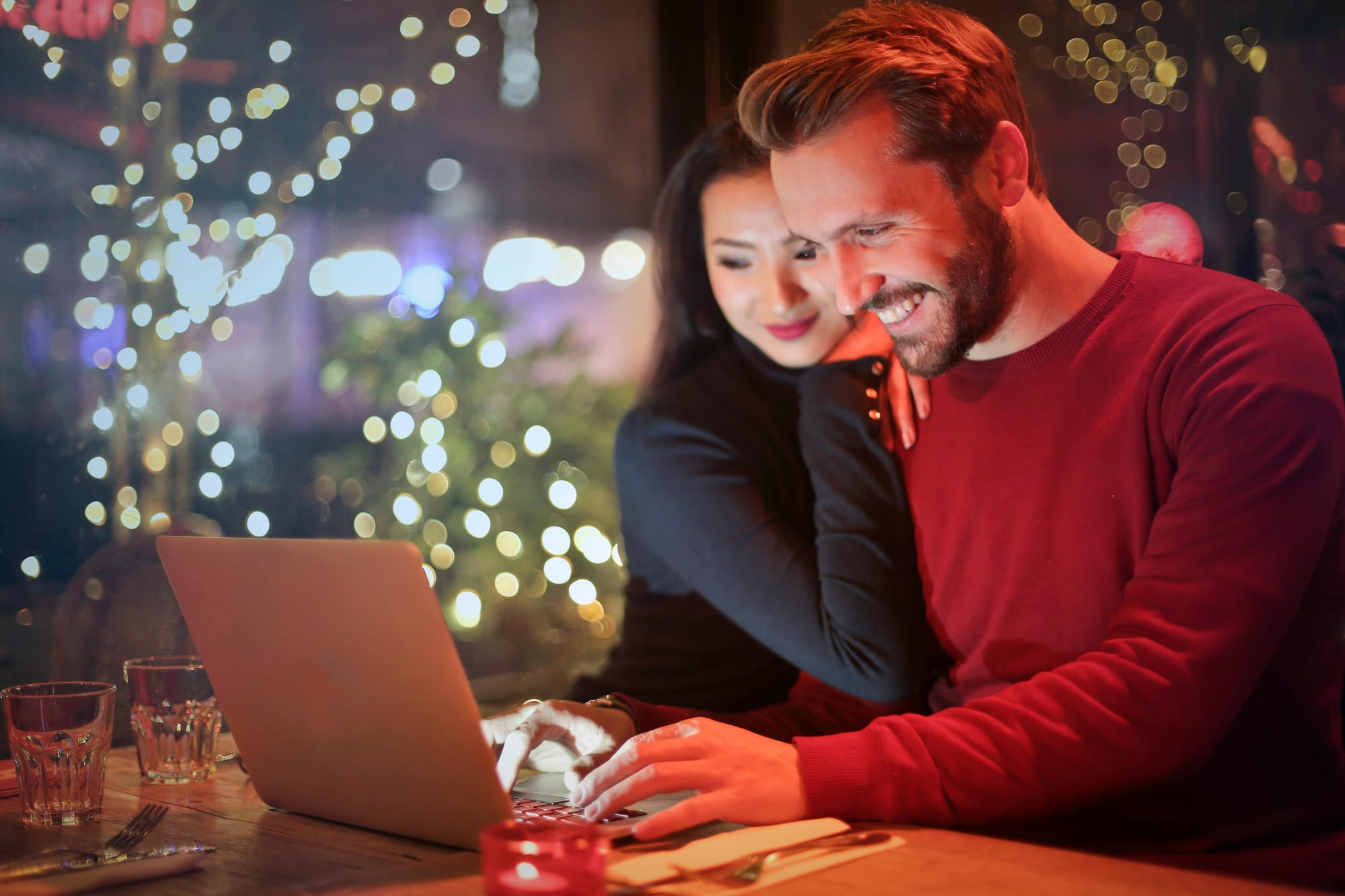 A man works with his laptop at a restaurant table, smiling, while a woman leans closely on his shoulder, smiling.