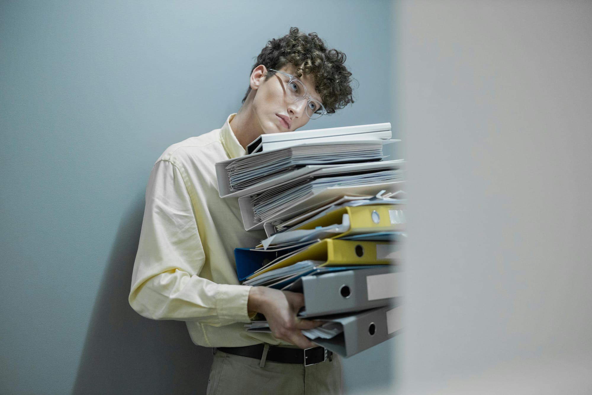 A man in office attire with glasses holds a large pile of files and paperwork, leaning his chin on the top in disdain.