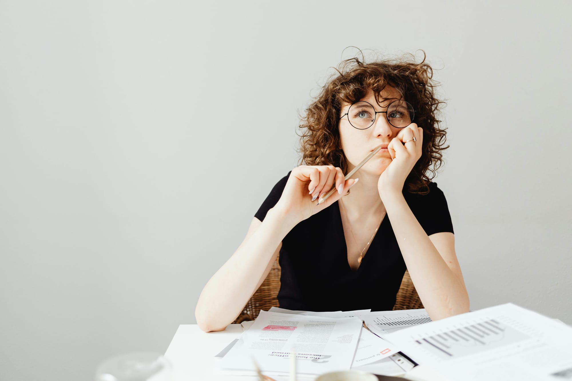 A curly-haired woman in thought, elbows leaning on a table filled with scattered paper.