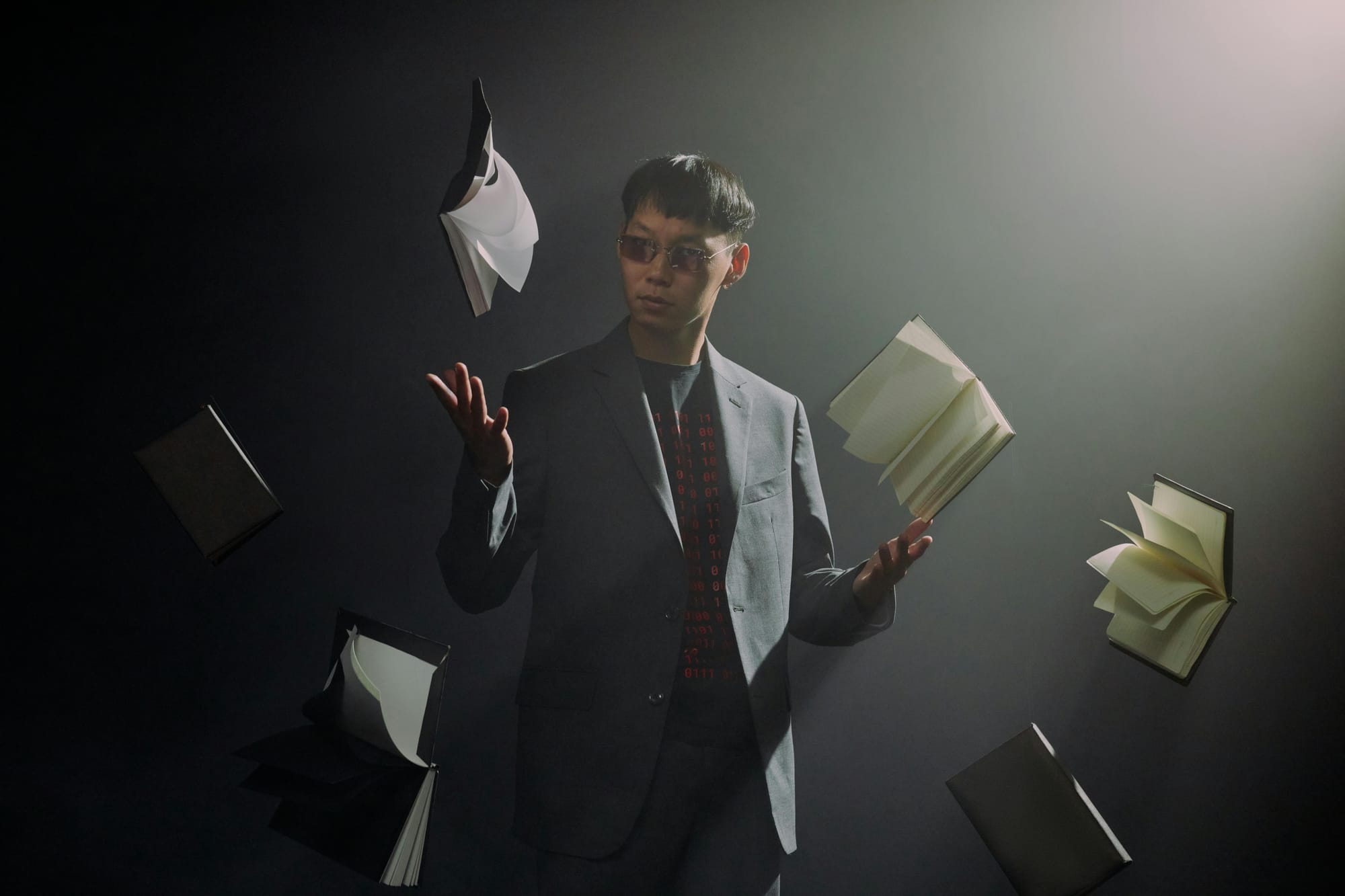 A suited man in red sunglasses looks to the camera as he seemingly controls floating, open books with his hands.