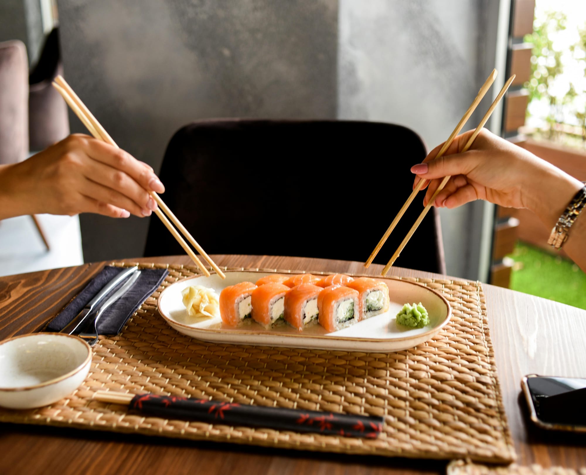 Two hands holding chopsticks approach one plate of sushi in the middle of the table.