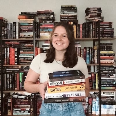 A woman stands in front of a bookshelf, holding a stack of books in front of her. She is wearing a light colored tshirt and jeans and has medium length brown hair.