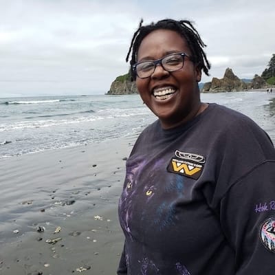 A woman with braids and glasses stands smiling on a beach. Her dark shirt has a Weyland-Yutani patch on it.