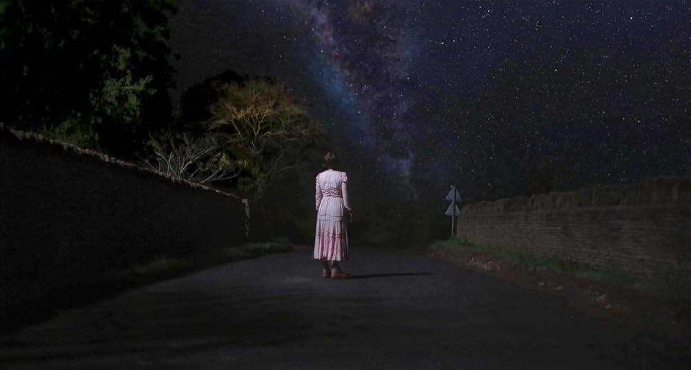 Jessie Buckley stands wearing a pink dress in a quiet country road, looking up at the bright stars in the night