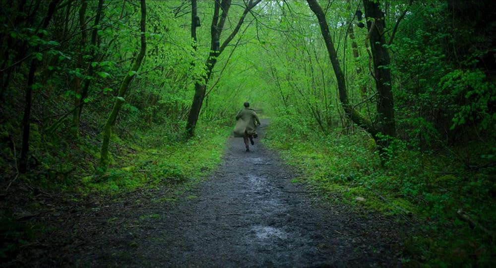 In a hyper green forest, Jessie Buckley runs down a path away from the camera