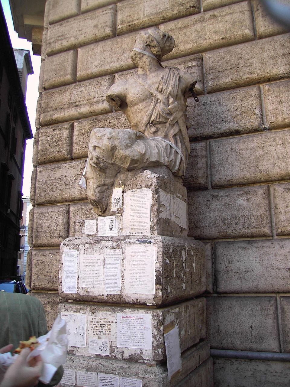 Photo of an ancient statue of a Greek warrior whose pedestal is covered with paper notices and pamphlets
