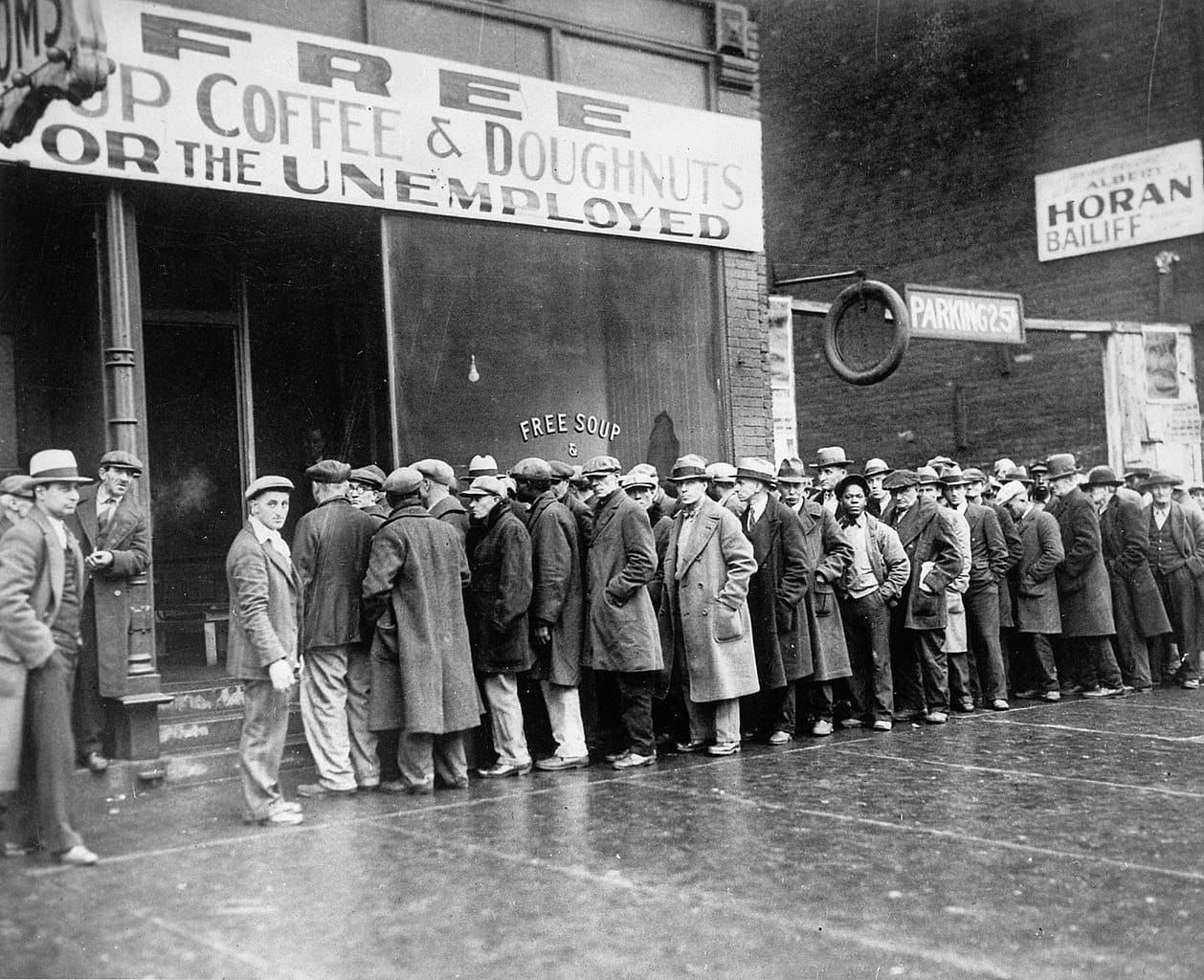 B&W photo of men queued before a storefront with a sign reading “Free Soup Coffee & Doughnuts for the Unemployed"