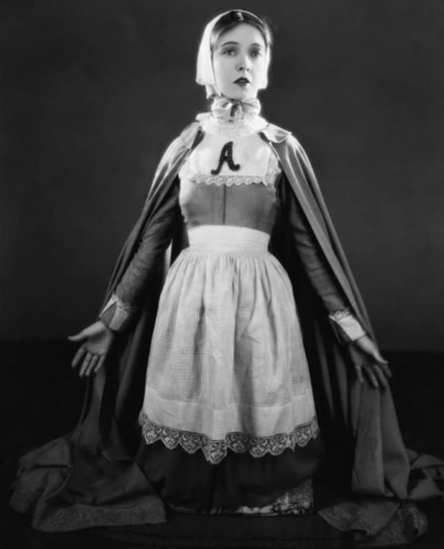 Black-and-white screenshot of a woman in Puritan dress with the letter “A” sewn onto her blouse