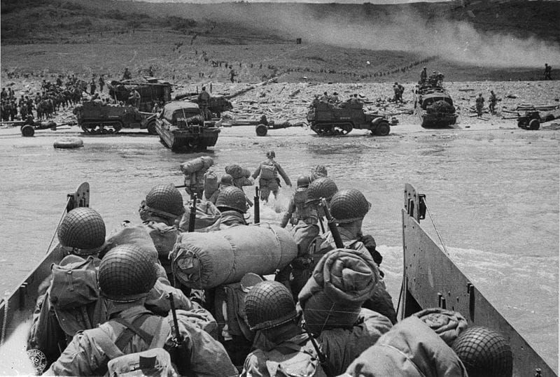 B&W photo of soldiers in a landing craft heading toward a beach filled with troops and military vehicles