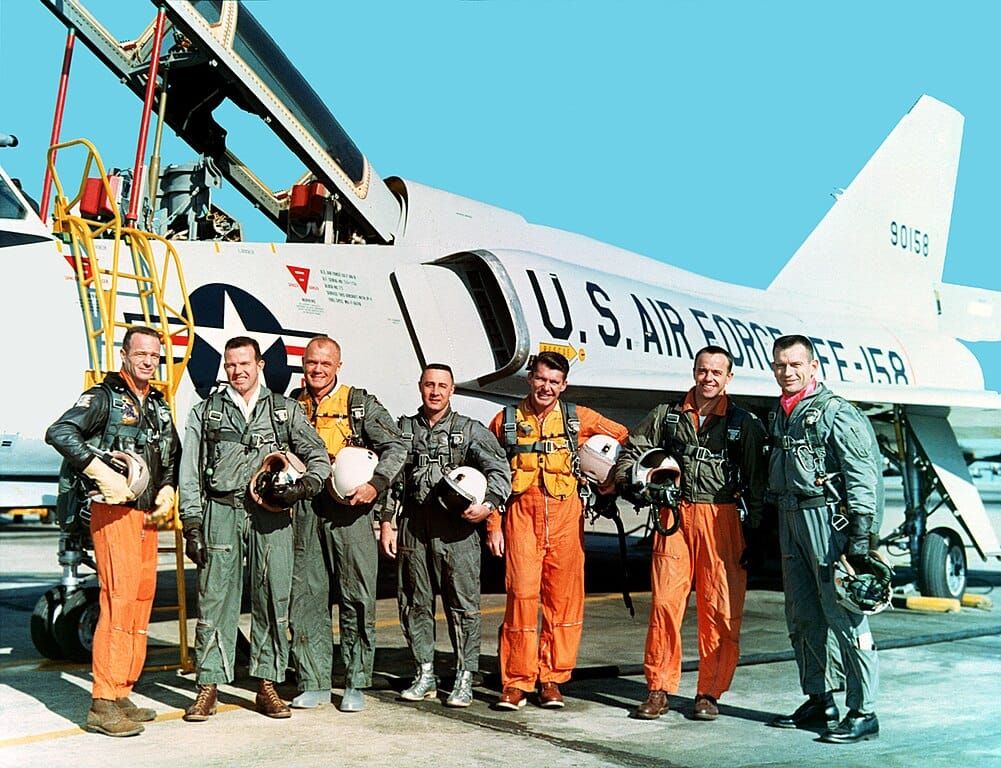 Photo of seven men in flight suits standing in front of a jet fighter