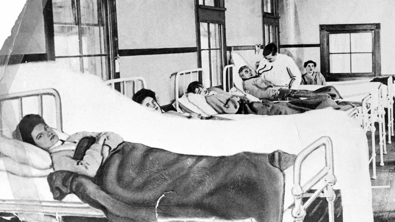 B&W photo of a woman lying on a bed in a hospital ward; behind her are four other patients and a doctor examining one