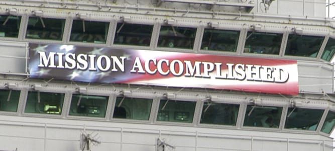 Banner reading “Mission Accomplished” against US flag background hanging from the superstructure of an aircraft carrier