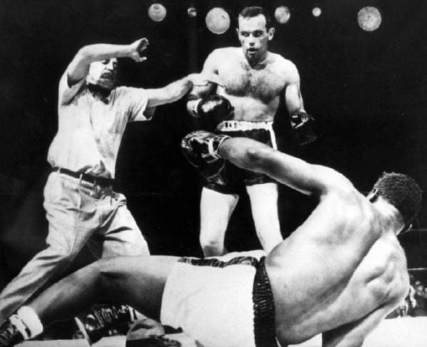 B&W photo of a referee intervening between two boxers, one of whom is lying on the ring floor