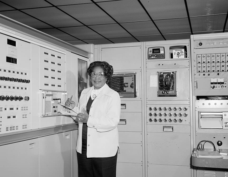 A Black woman standing in front of an electronic, mainframe computer