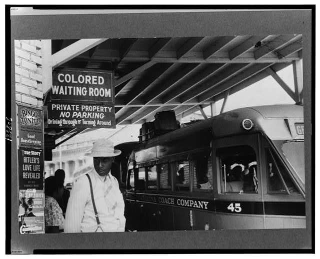 B&W photo of a Black man standing under a sign that reads, “Colored Waiting Room”; a bus is in the background
