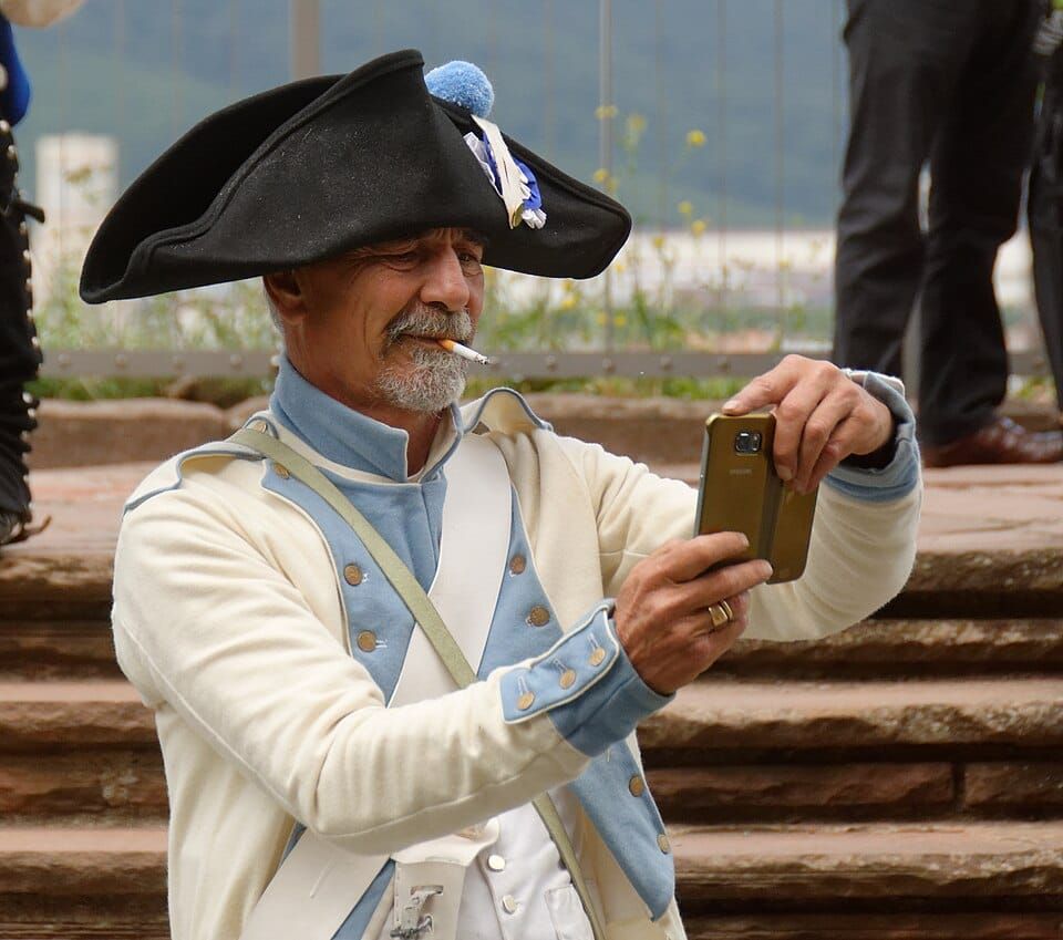 Photo of a man in 18th-century dress smoking a cigarette and taking a photo with with a smartphone